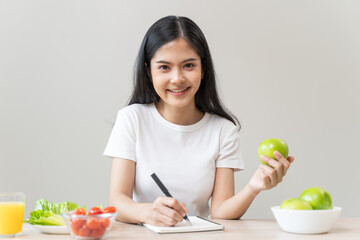 Asian beautiful woman i white t shirt writing note on the table and holding green apple with salad...