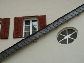 Light-colored building facade with rectangular and oval windows, red shutters, and slanted roof element. Copyspace.