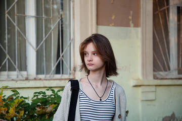 Young woman standing in front of building with shoulder bag and looking on summer outdoors city street. Cute natural teen girl 