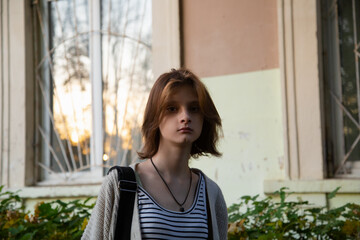 Young woman standing in front of building with shoulder bag and looking on summer outdoors city street. Cute natural teen girl