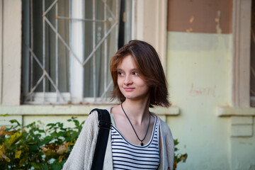 Young smiling happy woman standing in front of building with shoulder bag and looking on summer outdoors city street. Cute natural emotion teen girl