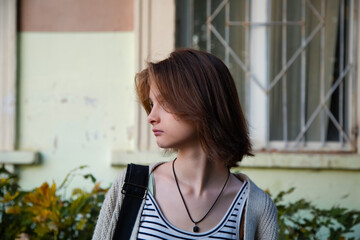 Young thinking woman standing in front of building with shoulder bag and looking on summer outdoors city street. Cute natural teen girl profile view closeup nature portrait