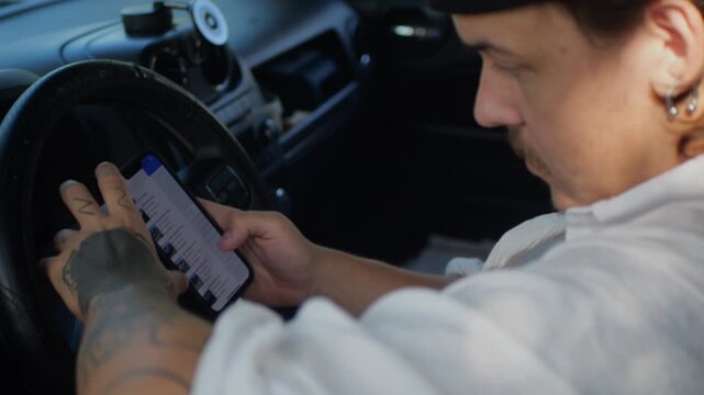 Man with tattooed hand in car using smartphone to browse online vehicle listings, concept of browsing car auctions, bidding or buying used cars via mobile app while sitting behind wheel