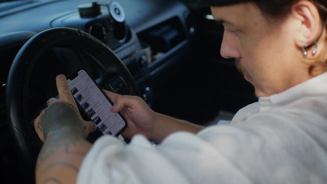 Man with tattooed hand in car using smartphone to browse online vehicle listings, concept of browsing car auctions, bidding or buying used cars via mobile app while sitting behind wheel