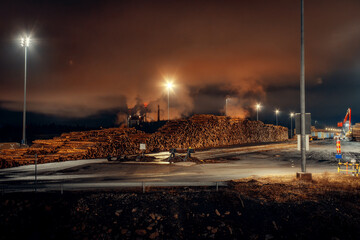 Nighttime industrial port landscape with large stacks of timber logs stored under artificial lighting. Floodlights illuminate wood cargo stockpiles and port infrastructure, while smoke and clouds 
