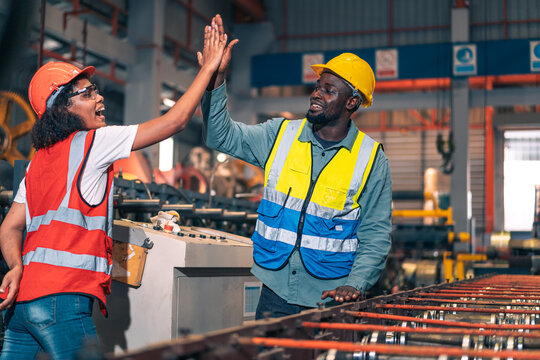African factory worker man woman shake hand high five success. Industrial engineer partner male female wear safety helmet vest in manufacturing plant. Heavy metal machine background. Happy diversity