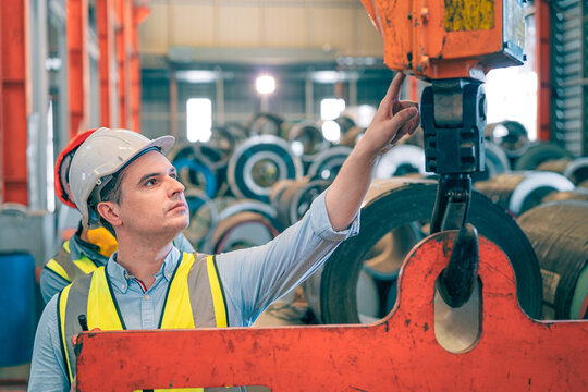 Caucasian industrial engineer men controlling overhead crane with remote in factory. Worker partner wear safety helmet vest lifting heavy metal coil material. Manufacturing hoist machine logistics