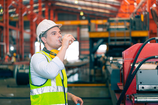 Thirsty Caucasian industrial engineer man wear safety helmet vest drinking water from plastic bottle in factory. Tired worker taking break refreshment. Manufacturing plant hydration health care