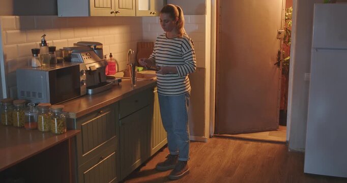 A young woman in casual clothes stacks table knives in a glass stand in a bright, modern kitchen in the evening. Everyday routine, lifestyle.