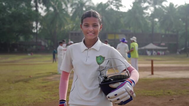 An Indian young, confident girl or female cricket batsman in a jersey and holding a helmet, stands outdoors on the ground, smiles looking at the camera, with teammates preparing in the background