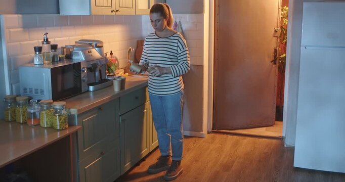 A young woman in casual clothes arranges paper cups on a countertop in a bright, modern kitchen in the evening. Everyday routine, lifestyle.