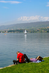 People relaxing on the grassy shore of Lake Viverone, Italy, with calm water, a sailboat, and snow-capped mountains beneath a clear blue sky.