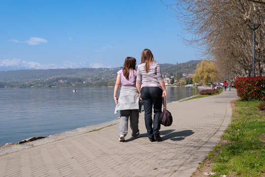 Two people walking along the promenade at Lake Viverone, Italy, enjoying a sunny spring day with calm water, mountains in the background, and a peaceful lakeside atmosphere.