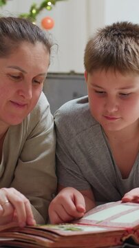 white mother and son studying holiday photos in album beneath christmas lights, quiet focus on pages, careful pointing and soft smiles, mother as family archivist preserving heritage