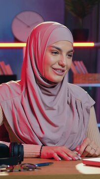Splendid in good mood carefree smiling young muslim woman in traditional hijab has pleasant video conversation with mate or relative when sitting at the table in beautifully decorated home office in