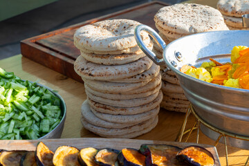 Tall stacks of pita bread beside bowls of chopped cucumber pickles and roasted eggplant on a wooden table at an outdoor cafe , ideal for Mediterranean cuisine catering and buffet concepts