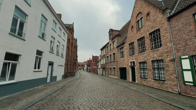 Scenic views of cobblestone streets in Bruges, Belgium, featuring traditional Flemish brick architecture, shops, and tourists walking past parked cars during a cloudy day in the historic center.

