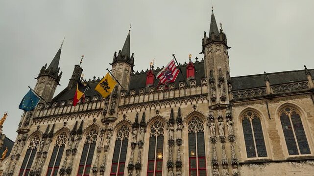 A detailed view of the Provincial Court on the Market Square in Bruges, Belgium. The neo-gothic building features flags waving and intricate stone details on a cloudy day.

