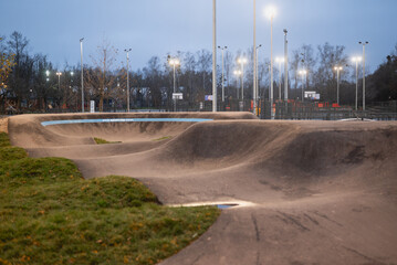 Modern asphalt pump track park for bicycles and skateboards illuminated by bright floodlights at twilight.