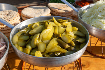 Metal bowl filled with sliced pickled cucumbers in brine on a wooden counter beside shredded cabbage and pita stacks at a casual cafe, ready as tangy burger toppings during lunch service