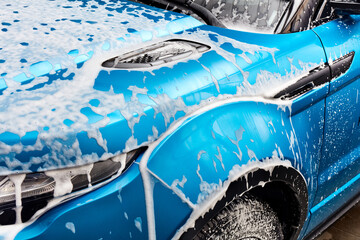 A bright turquoise car is being washed. Thick white foam covers the hood, headlight, fender, and tire. Soap is dripping down the side of the clean automobile.
