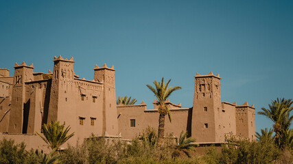 Ancient earthen kasbah rises above palm trees under a clear blue sky, showcasing traditional Moroccan architecture in warm desert light.