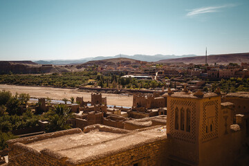 A&iuml;t Ben Haddou, Morocco. Panoramic view of the UNESCO-listed ksar with adobe towers, palm-lined riverbed and desert town stretching toward the Atlas Mountains under clear sky.