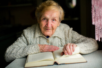 Elderly woman flips through a book at her table, warm light highlighting her gentle expression and cozy knitted sweater.