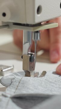 closeup sewing machine needle stitching grey fabric, hands guiding seam under presser foot in bright studio light, precision topstitching on durable knit, focused workshop environment, garment