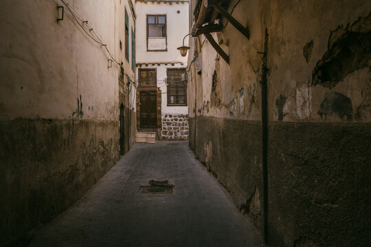 A quiet narrow alley in the old city of Damascus, lined with weathered stone walls, traditional houses, and soft natural light reflecting the city&rsquo;s historic character.