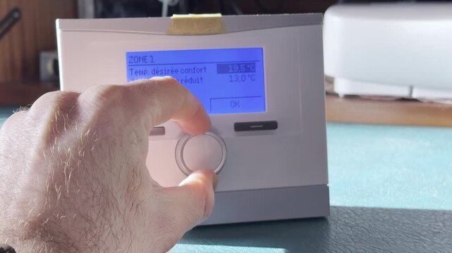 Male hand adjusting digital room thermostat with blue backlit LCD on desk, setting comfort temperature for home heating, HVAC control, energy efficiency concept, with french text