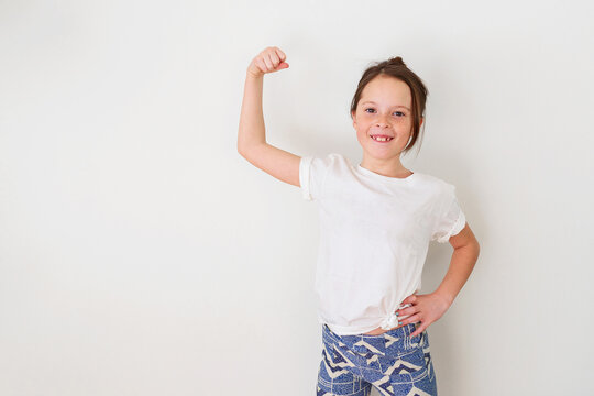 Portrait of a young smiling girl in a white t-shirt standing in front of a white wall flexing her muscles