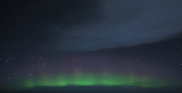 Low band of green and faint magenta aurora glowing along a distant horizon beneath heavy clouds, casting a subtle line of color across the brooding darkness of the polar night

