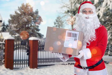 Obraz premium Santa Claus holding parcel near gate and fence on snowy street, outdoors