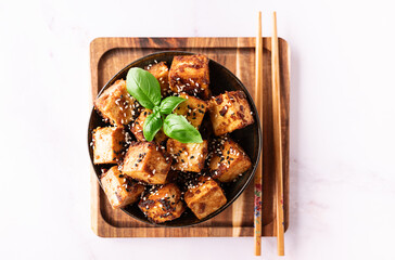 Fried tofu with sesame seeds, soy sauce, spices decorated with fresh basil leaves on white marble table top view. Healthy Asian food concept. Vegan food.