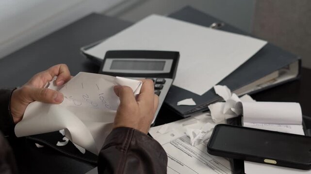 Frustrated man tearing debt papers at his desk, surrounded by calculator, laptop, and document folders, representing financial problems, anger, and economic pressure.
