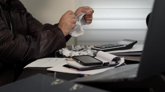 Young man tearing and crumpling debt papers at his desk, showing anger and frustration over unpaid bills, with calculator, laptop, and document folders visible in the background.