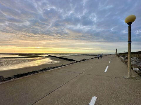 Two people walking on seaside promenade at sunset in Berck, France, along the Authie Bay, overlooking low tide sand flats and tidal channels, dramatic cloudy sky, coastal tranquility - Powered by Adobe