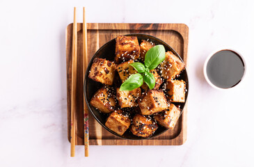 Fried tofu with sesame seeds, soy sauce, spices decorated with fresh basil leaves on white marble table top view. Healthy Asian food concept. Vegan food.