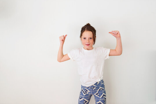 Portrait of a young smiling girl in a white t-shirt standing in front of a white wall flexing her muscles