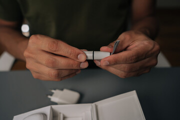 Obraz premium Close-up cropped shot of unrecognizable man sitting at table unpacking new electronic cigarette from box, holding heating element and cover. Concept of modern alternative to traditional smoking.