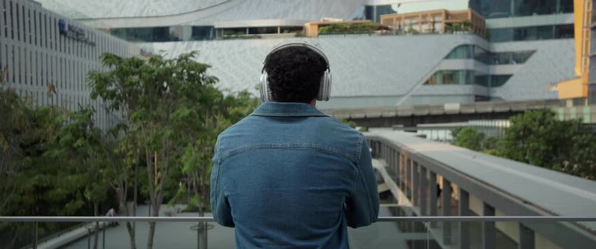 Hispanic man wearing denim jacket listens to music via earphones by railing in city anamorphic lens. Latin male with headphone stands on terrace and looks at business center