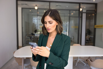 Serious beautiful Hispanic executive woman typing on smartphone, standing in office, using...