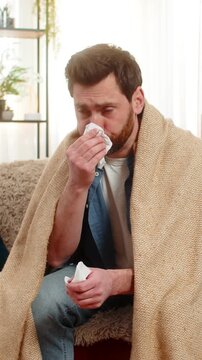 Young sick ill man at home sits on the sofa, wrapped in blanket, sneezing and quickly reaching for tissues. Caucasian guy wipes his nose, looking unwell, possibly suffering from a cold or flu symptoms