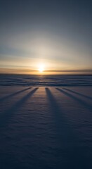 Minimalist winter landscape showing long shadows on a frozen snow-covered lake during a golden sunset with a clear sky.
