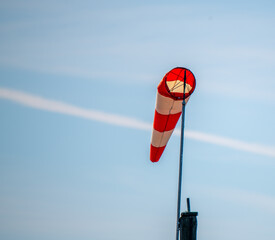 An orange and white windsock silhouetted against a blue sky and a jet contrail. 