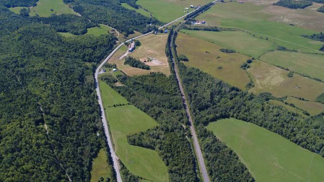 High-altitude aerial view of a small rural village showing winding roads, houses, a railway line, agricultural fields, and the surrounding forest. Padoue, Quebec, Canada, 2025.