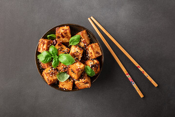 Fried tofu with sesame seeds, soy sauce, spices decorated with fresh basil leaves on black stone background top view. Healthy Asian food concept. Vegan food.