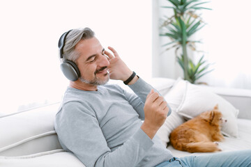 Mature man relaxes on sofa wearing wireless headphones while enjoying music at home. His peaceful...