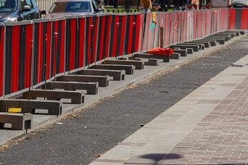 Construction barriers in bright red line a street indicating ongoing development. Pedestrians navigate around the site while cars move in the background